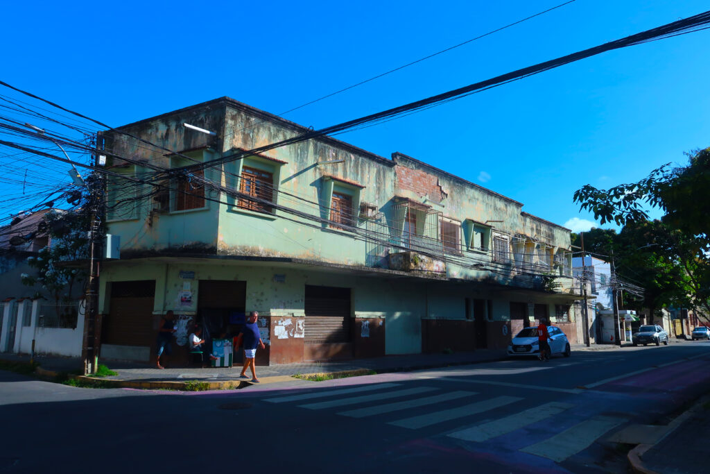 A imagem mostra um prédio de esquina de dois andares em uma área urbana sob um céu azul. O prédio está desgastado e precisa de reparos, com pintura desbotada e manchas de mofo. As janelas têm grades de proteção, algumas danificadas. Na calçada, pessoas caminham e conversam, e um carro está estacionado na rua. Cabos elétricos são visíveis ao longo da imagem, destacando a infraestrutura elétrica. A rua tem faixas de pedestres, e há árvores na calçada, adicionando um pouco de verde ao cenário urbano.