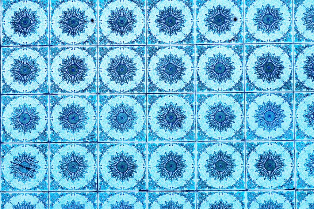 A imagem mostra um padrão de azulejos azuis com desenhos circulares complexos no centro de cada um. Os círculos centrais lembram mandalas ou flores estilizadas, com detalhes intricados em várias tonalidades de azul. Cada azulejo é enquadrado por arabescos nos cantos.