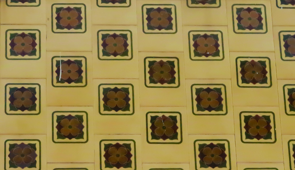 A imagem mostra um padrão de azulejos amarelos, cada um decorado com um motivo floral central marrom e verde, dentro de um quadrado. Os azulejos são dispostos de forma regular e simétrica.