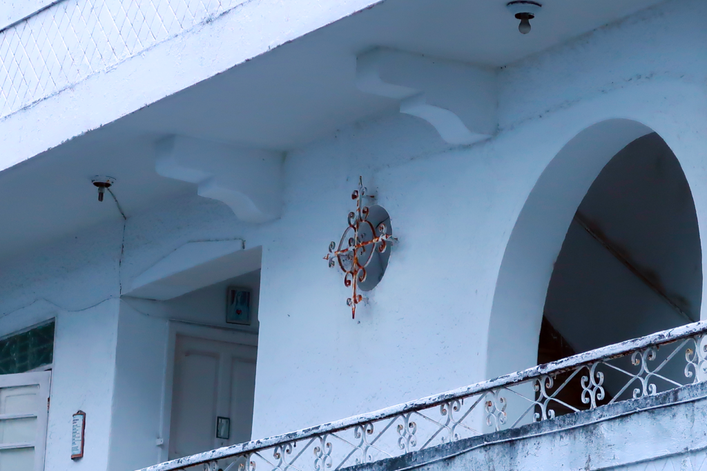 A imagem mostra a fachada de um edifício branco com detalhes arquitetônicos simples. Há um terraço com grade de metal decorativa que parece enferrujada. Na parede, há um ornamento de metal enferrujado em formato de cruz decorativa.