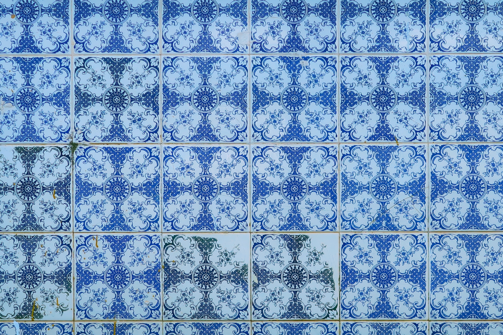 A imagem mostra um padrão de azulejos azuis e brancos com desenhos intrincados. Cada azulejo possui um círculo central com motivos florais e arabescos, formando um padrão repetitivo e simétrico. Os azulejos apresentam leves variações de cor e alguns sinais de desgaste.