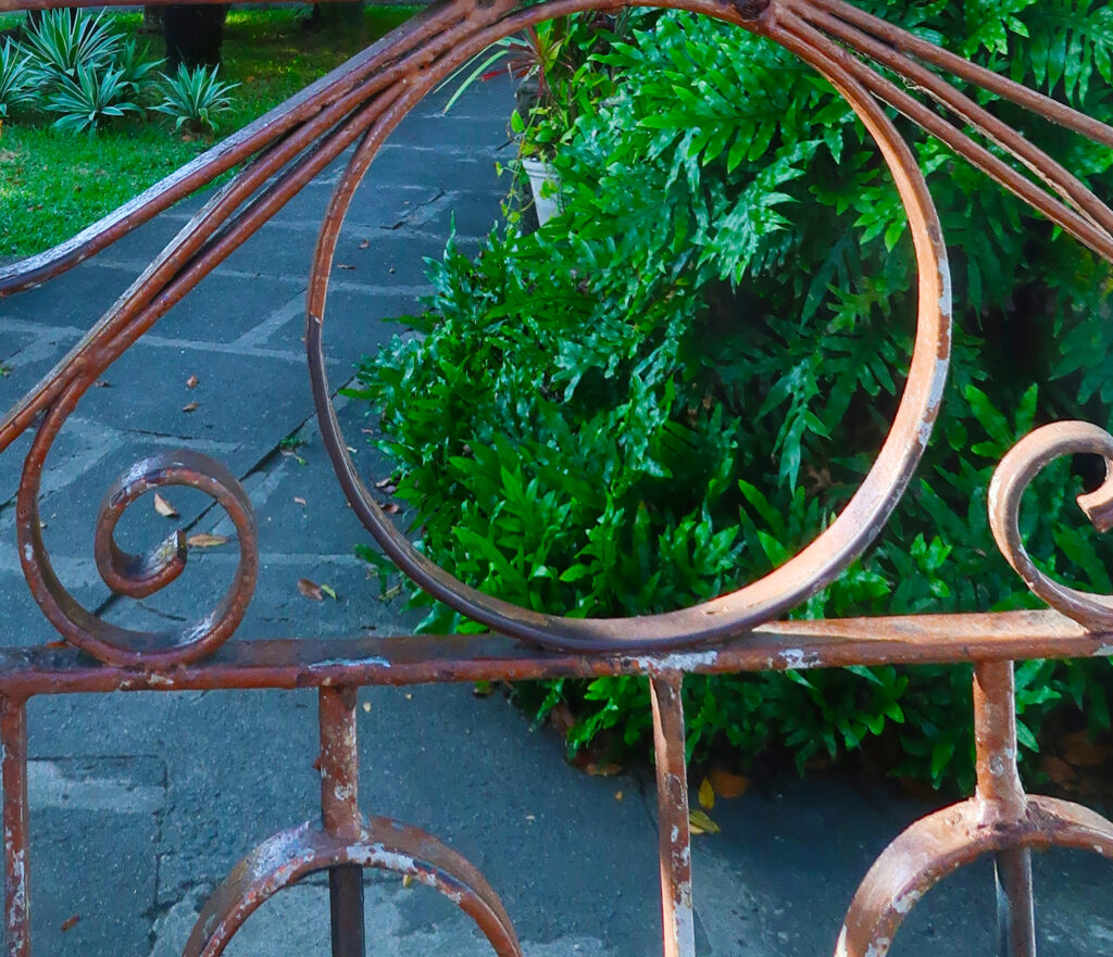 A imagem mostra um portão de metal com um design decorativo enferrujado, com padrões de espirais. Ao fundo, há um caminho de pedra cercado por vegetação verde, sugerindo um jardim ou quintal bem cuidado.