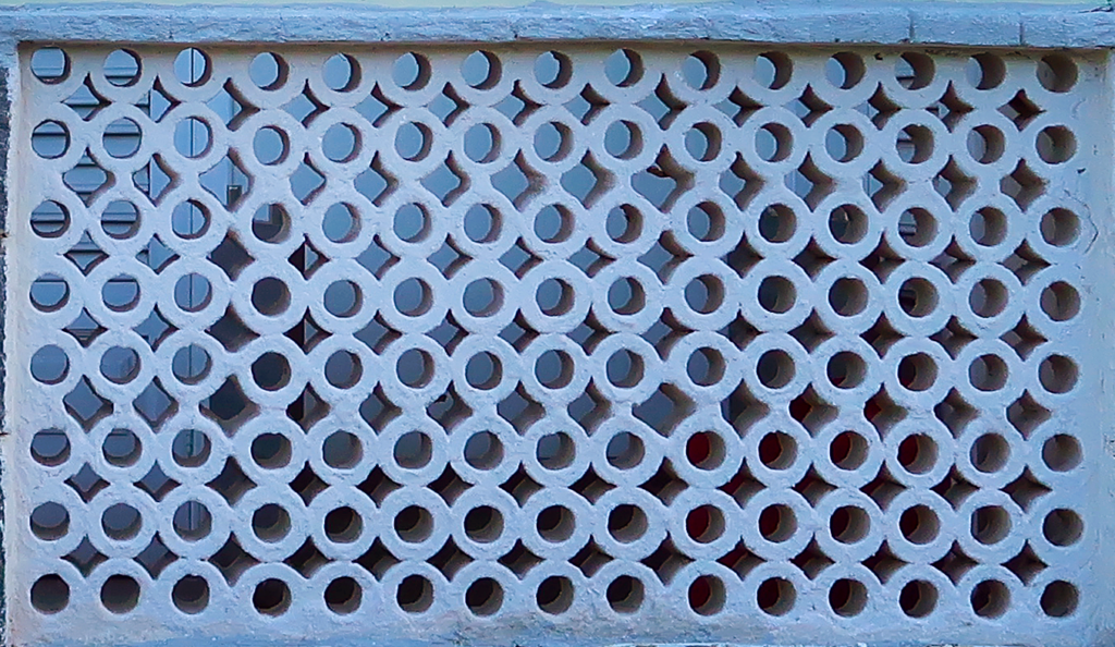 A imagem mostra uma parede de elementos vazados de concreto com um padrão repetitivo de círculos e losangos. Esse tipo de elemento arquitetônico é utilizado para permitir a ventilação e a passagem de luz, enquanto ainda mantém a privacidade e a segurança.